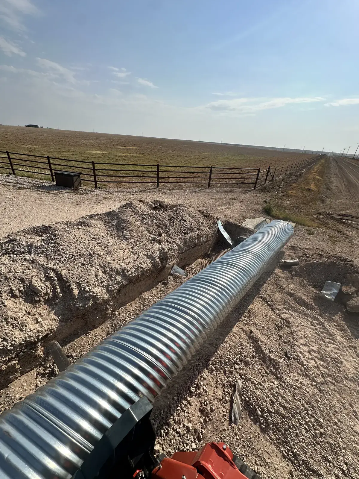A long galvanized culvert pipe laid in a trench running toward a ranch gate on the Texas Panhandle plains