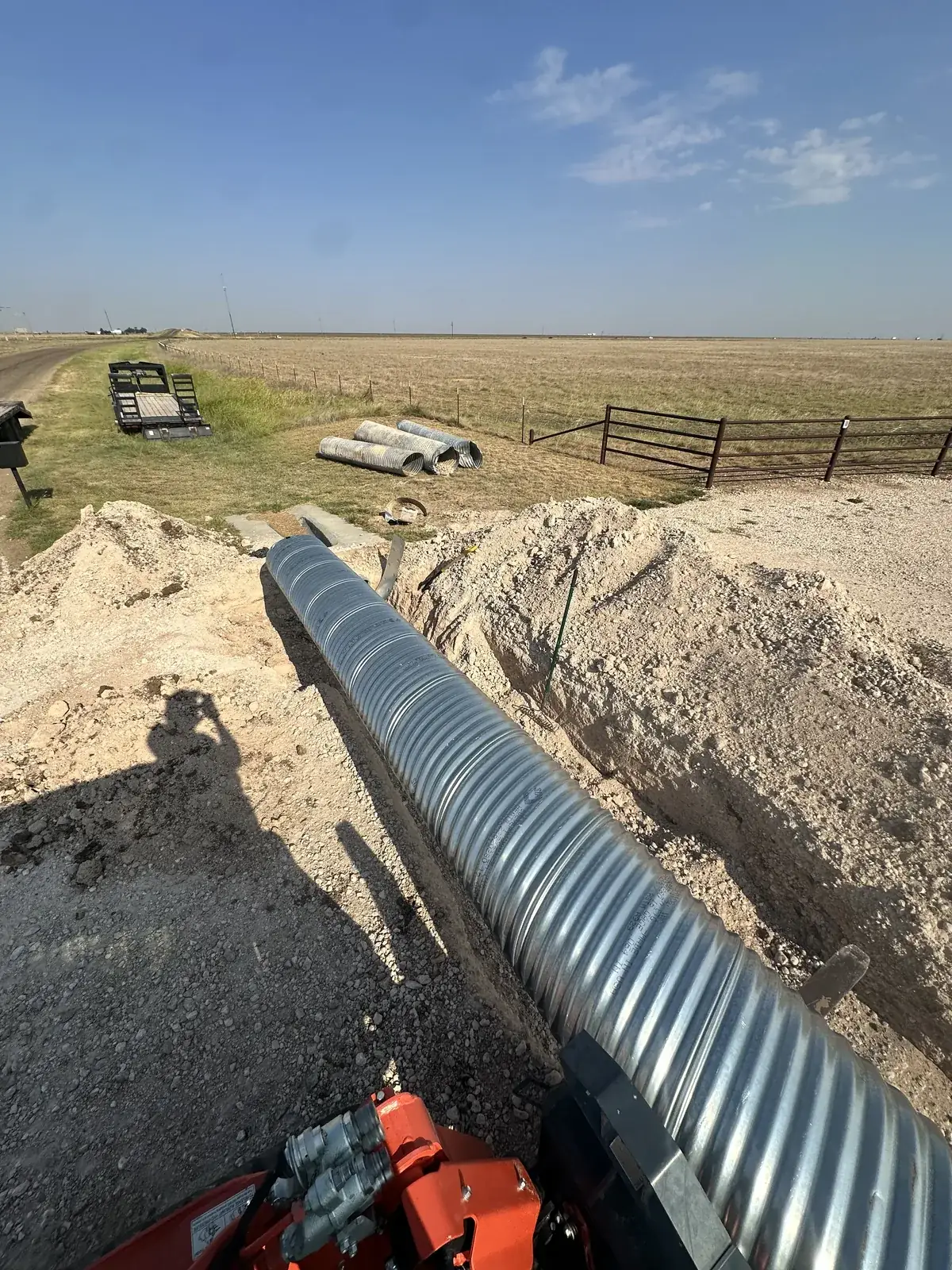 A galvanized corrugated-metal culvert pipe being set into an excavated trench by a loader at a Texas Panhandle ranch entrance
