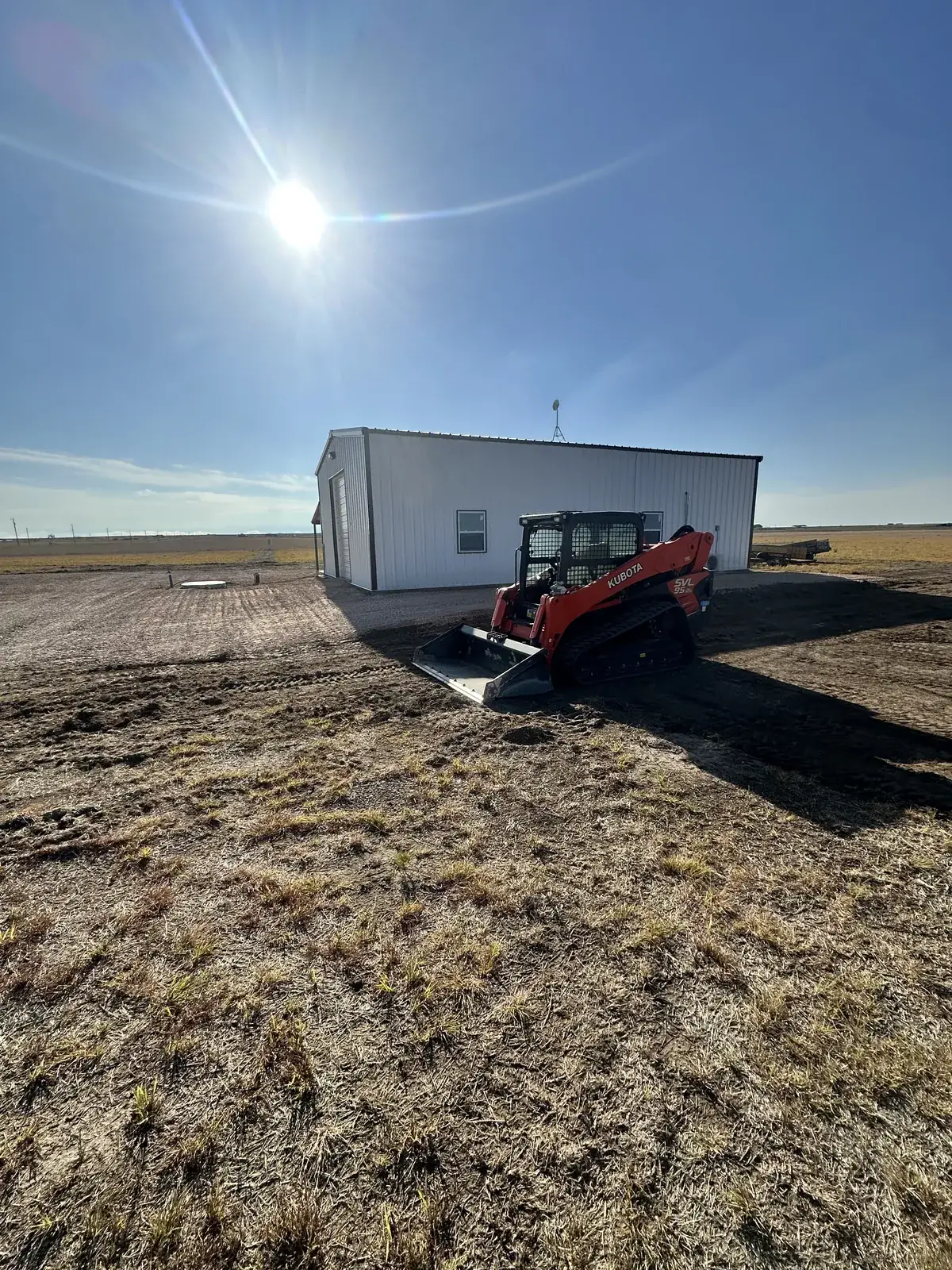 A Kubota compact track loader parked at a rural job site beside a new metal building in the Texas Panhandle